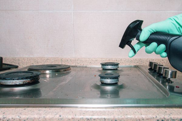 A person wearing green gloves is using a spray bottle to clean a stovetop in a kitchen. The surface is slightly dirty, and the cleaner is being applied for better hygiene.